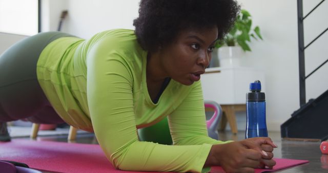 Young African-American woman in yellow long-sleeved shirt performing plank exercise indoors. The setting includes a pink yoga mat, workout equipment, and a water bottle. Perfect for illustrating home fitness, determination in workouts, or promoting active lifestyles. Use in wellness blogs, fitness apps, or health magazine articles.