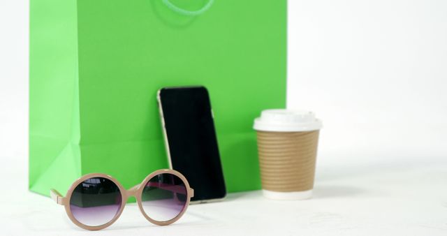 Sunglasses and a smartphone lean against a large green shopping bag, suggesting a minimalist lifestyle and fashion sense. Beside them, takeaway coffee emphasizes a busy, upbeat routine. Perfect for blogs about minimalism, lifestyle tips, shopping, or fashion items. Great for illustrating trendy, on-the-go contemporary lifestyles.