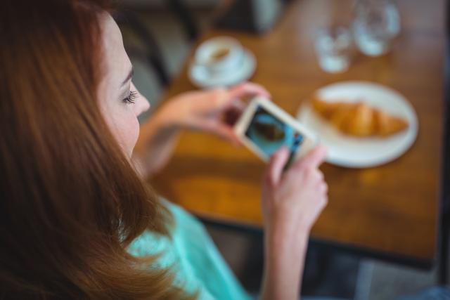Woman clicking photo of croissant from mobile phone in cafÃ©