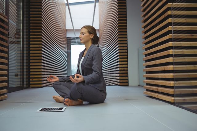 Businesswoman practicing yoga in a modern office corridor. She is seated in a cross-legged position, meditating with eyes closed. This image can be used for promoting corporate wellness programs, stress relief techniques, mindfulness in the workplace, and healthy lifestyle choices for professionals.