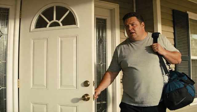 Adult man entering sunlit suburban home carrying navy duffel bag, looking concerned