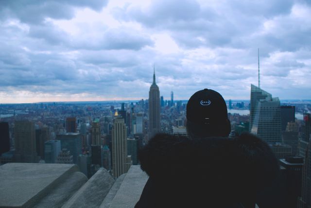 Person standing on tall building terrace overlooking New York City skyline with iconic skyscrapers and cloudy sky. Could be used to illustrate travel, urban exploration, and adventure themes, or for promoting tourism in New York.