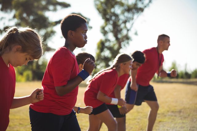 Trainer leading a group of kids through exercises in an outdoor boot camp on a sunny day. Majority of the focus is on the group, showing teamwork and physical activity. The image can be used in articles or advertisements about fitness, children's health, outdoor activities, summer camps, sports training, and group exercise programs.
