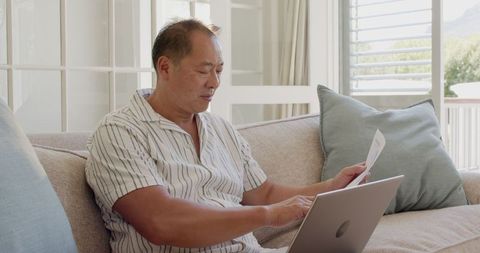 Senior Man on Sofa Using Laptop and Reviewing Papers at Home