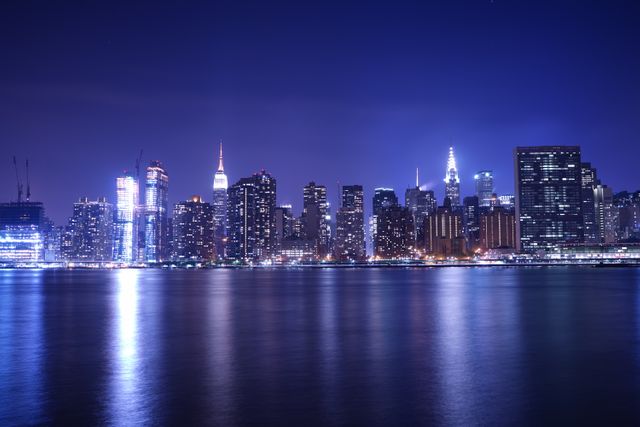 This image captures the iconic Manhattan skyline across the East River at night, showcasing illuminated skyscrapers and their reflections in the water. Ideal for use in travel blogs, tourism brochures, and architectural showcases to evoke a sense of urban sophistication and vibrant city life.