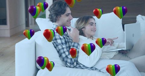 Cuddling couple relaxing on couch watching laptop and smiling with floating rainbow hearts