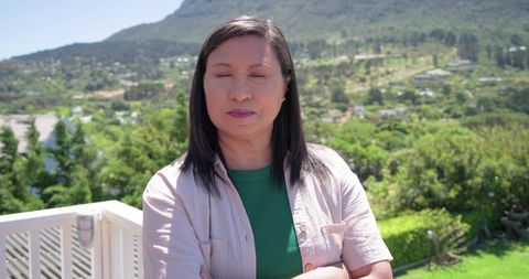 Mature asian woman standing on white balcony crossing arms overlooking sunlit green valley