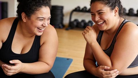 Two friends laughing after workout on blue mat in gym, celebrating progress together