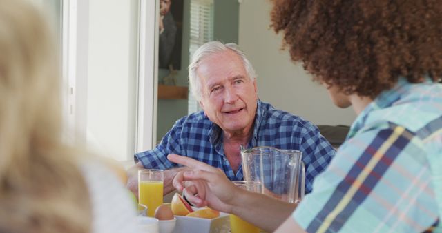 Elderly man with white hair and a plaid shirt engaging in conversation with young adults over breakfast, featuring glasses of orange juice and a casual dining setting. Ideal for themes of family bonding, intergenerational interaction, and domestic life.