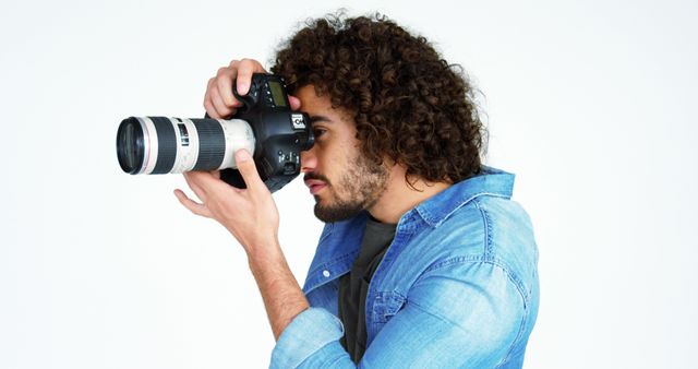 Young man with curly hair holding DSLR camera capturing photos, against neutral white background. Ideal for use in articles about photography tips, equipment reviews, or creative careers. Can be used in promotional materials for camera brands or visual arts workshops.