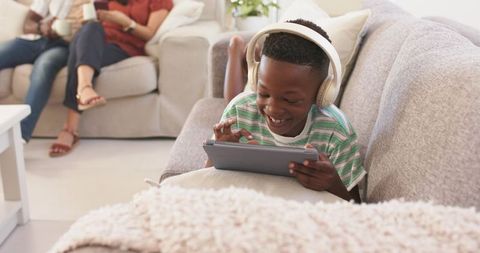Smiling boy wearing over-ear headphones using tablet on cozy living room sofa