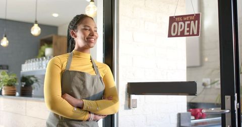 Smiling barista greeting customers at open café door in mustard turtleneck and gray apron