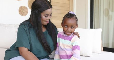 Warm african american mother comforting smiling daughter on sofa in cozy indoor pastel home