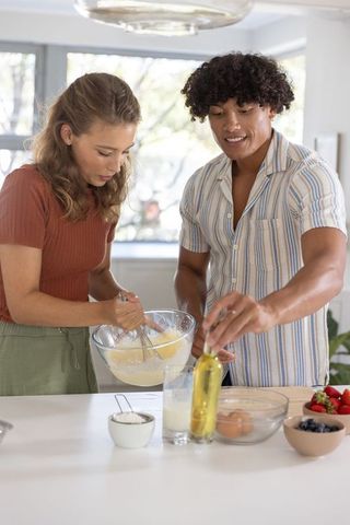 Young couple baking together at kitchen island, whisking batter and adding oil
