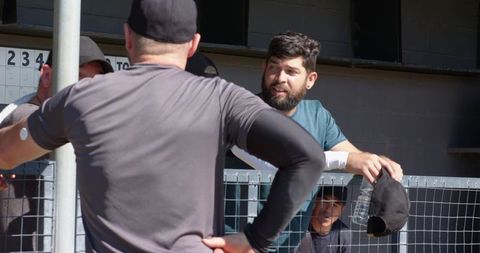 Sunlit dugout diverse male teammates leaning railing holding cap and water bottle