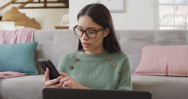 Young woman sitting on floor using smartphone and laptop in home living room. Can use for concepts of technology, remote work, multitasking, and modern lifestyle.