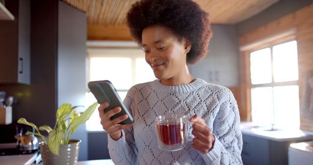 Happy african american woman with afro holding cup of tea and using smartphone in sunny kitchen. Lifestyle, free time, communication and domestic life, unaltered.