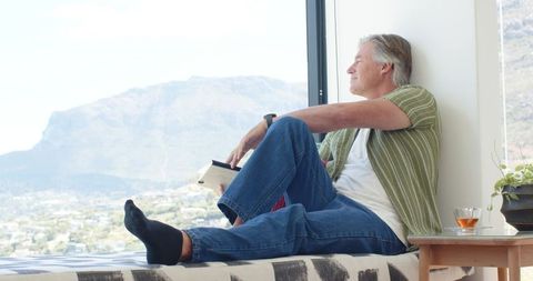 Senior man relaxing on window bench reading book gazing at panoramic mountain valley view