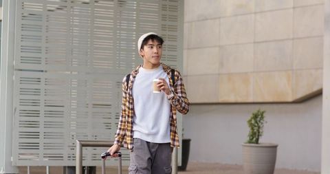 Young Asian Man Walking with Carry-On Luggage and Coffee Wearing Beanie and Plaid Shirt