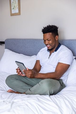 African american man sitting cross-legged on bed using tablet in cozy modern bedroom