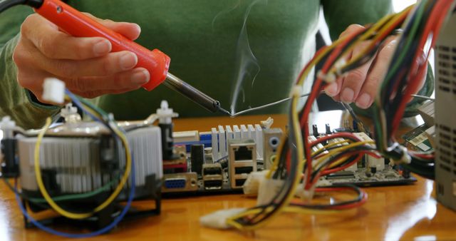 A person is soldering a component on an electronic board, with copy space. Various electronic parts and tools are laid out on a wooden table, indicating a work environment for an electronics technician or hobbyist.