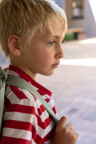 Blonde boy carrying backpack walking through sunlit school courtyard looking thoughtful