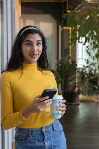 Young indian woman in yellow top holding smartphone and coffee in modern coworking space