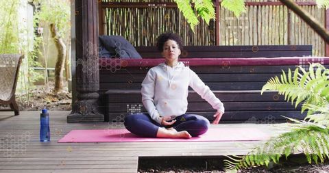 Woman meditating on pink yoga mat on covered deck surrounded by lush garden greenery