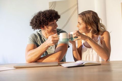 Sunlit diverse couple toasting with matching mugs at home desk, smiling and connecting