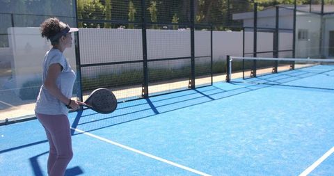 Woman standing with padel paddle on sunlit blue court preparing to serve