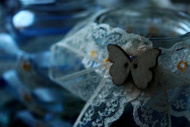 Close-up of a jar adorned with delicate lace and a butterfly ornament for a rustic feel. The background showcases more jars, adding to the cozy yet elegant atmosphere. Ideal for use in wedding invitations, DIY craft blogs, or rustic home decor inspiration.