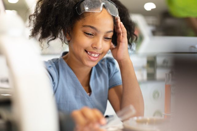 Smiling biracial elementary schoolgirl with head in hand performing chemistry practical in lab. unaltered, education, laboratory, stem, scientific experiment, protection and school concept.