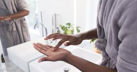 African american man applying moisturizer at modern bathroom vanity in grey bathrobe
