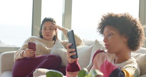 Multicultural women relaxing on sofa, scrolling smartphones in sunlit living room