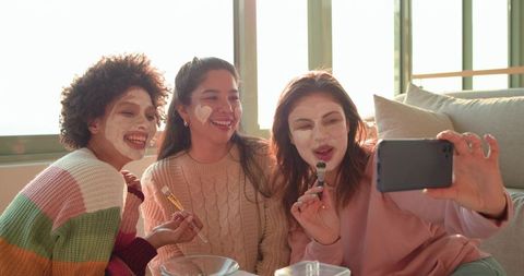 Diverse friends enjoying skincare night taking selfie on sunlit sofa