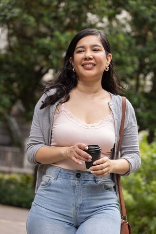 Smiling woman walking holding coffee to-go cup in urban park wearing cardigan and jeans