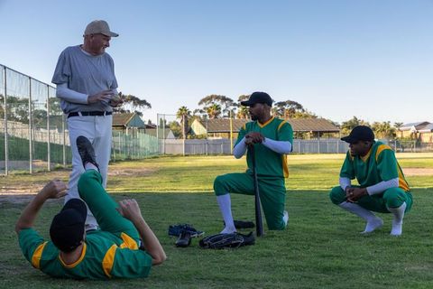 Coach directing diverse baseball team practicing on sunlit community field bat drills