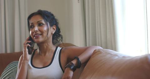 Relaxing woman talking on smartphone on tan sofa wearing white tank and bracelets