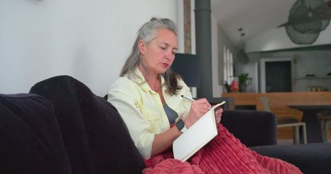Senior woman journaling on sofa with smartwatch and red throw in open-plan living