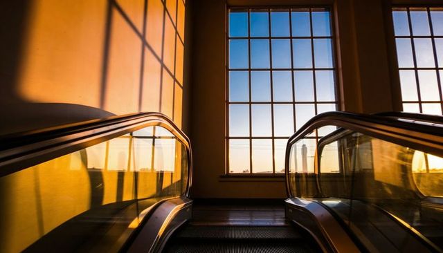 Golden-hour escalators leading to grid window in modern rail station interior