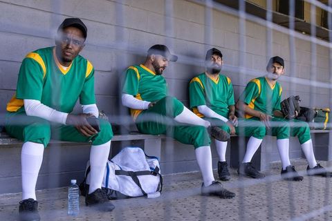 Baseball players sitting in dugout checking phone and waiting to play in green uniforms