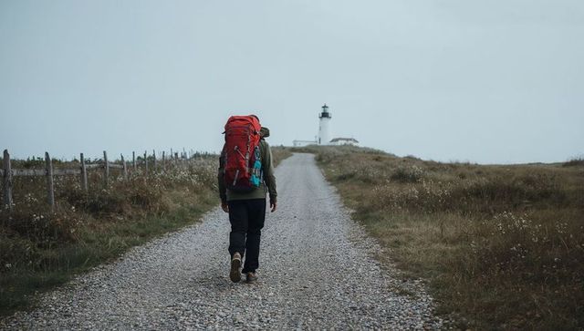 Solo hiker walking gravel coastline path toward distant lighthouse carrying red backpack