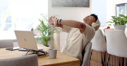 Young man stretching at home desk with laptop, headphones, coffee and green plants