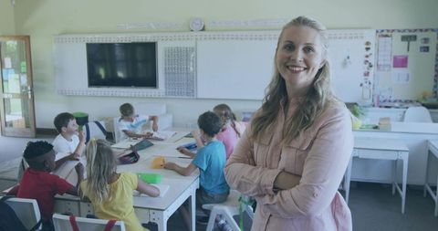 Smiling teacher standing with arms crossed watching elementary students learning with tablet