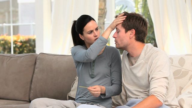 Woman in casual attire tenderly touches her husband's forehead, checking his temperature with a thermometer in hand, while seated on a couch in a cozy living room. This image captures a moment of care and concern between partners, highlighting themes of love and support during illness. This could be used in articles about healthcare, relationship advice, or stress management.