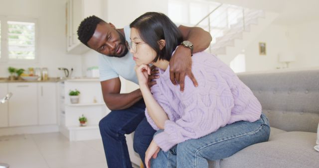 Sad diverse couple sitting on couch and talking in living room. Spending quality time at home concept.