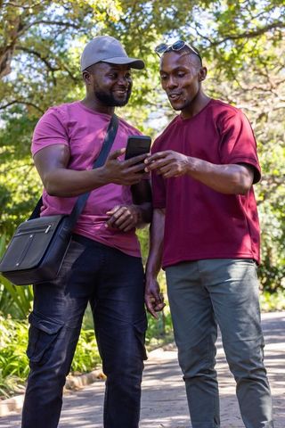 Friends sharing smartphone while walking under sunlit park trees
