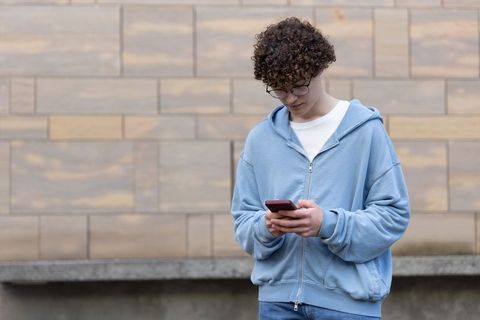 Teen male standing outdoors holding smartphone wearing blue hoodie and jeans by stone wall