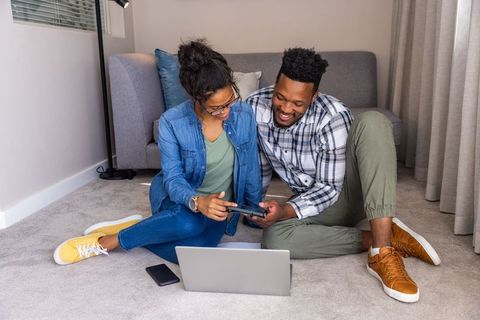Couple sitting on living room floor sharing smartphone while using laptop
