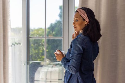 Woman standing beside sliding glass door holding cup touching face wearing denim shirt